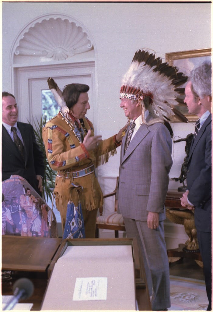 Photo of a man in deerskin clothing talking with Jimmy Carter, who is wearing a Native-American headdress. 