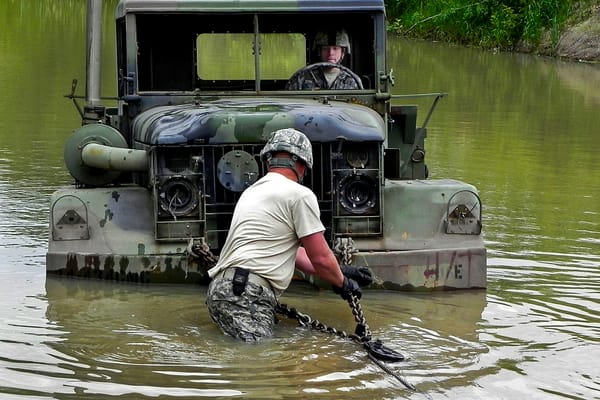Photo of a soldier attaching a tow chain to a truck that is stuck in the middle of a river