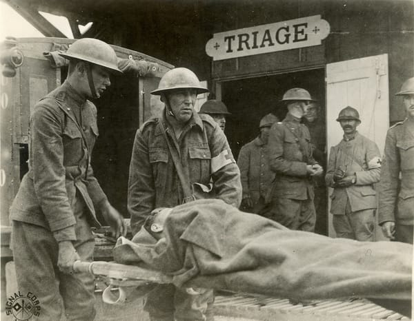 B&W photo of two US Army medics carrying a wounded soldier into an aid station. US and French soldiers are in the background