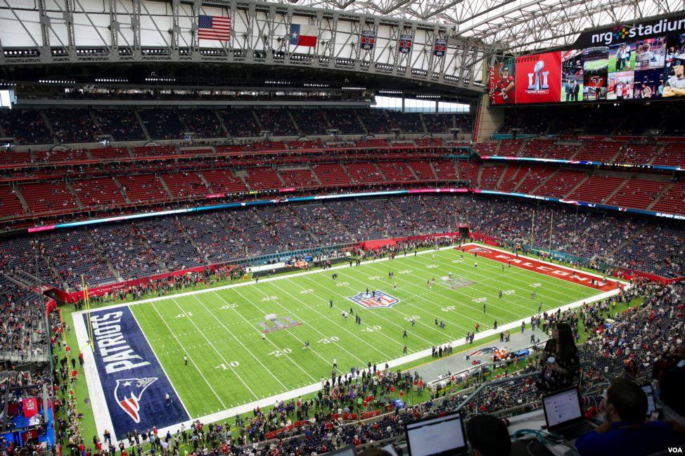 Color photograph of the interior of a large football stadium; the logo of Super Bowl LI is displayed on the scoreboard