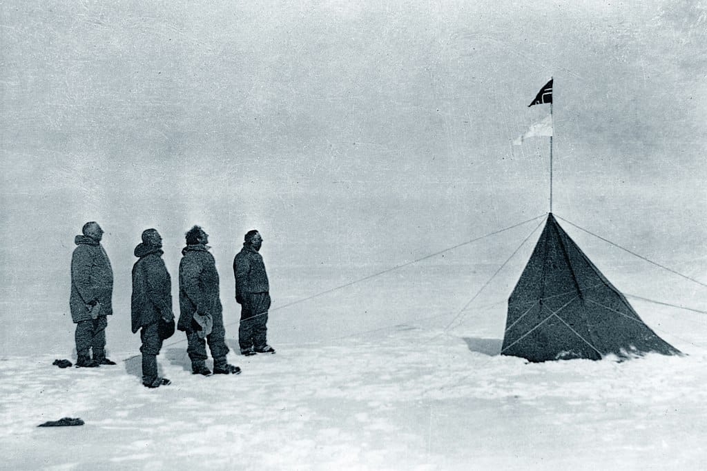 Black-and-white photo of four men standing in the snow and looking at a small, Norwegian flag flying from a pole