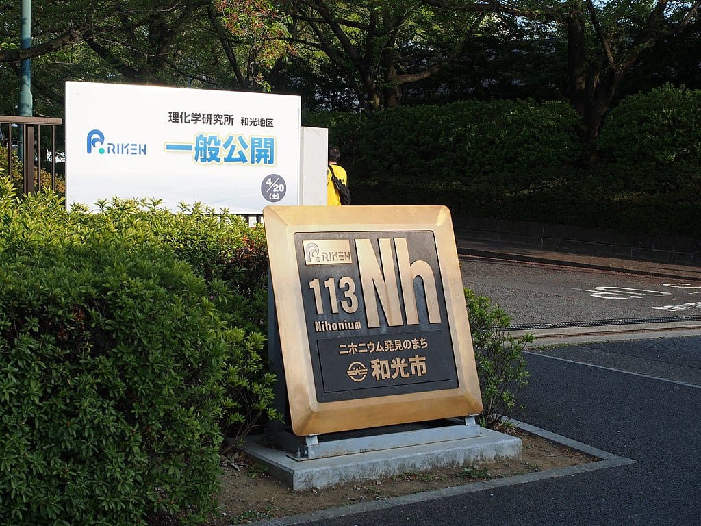 Marker bearing the periodic table entry for element 113 next to a clump of bushes and a sign for the Riken institute.