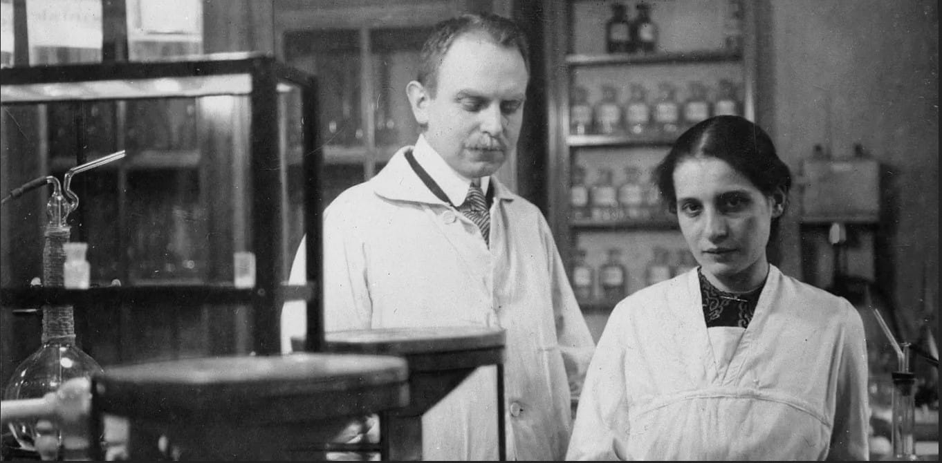 Black-and-white photo of a man and a woman, wearing lab coats, standing in a chemistry laboratory