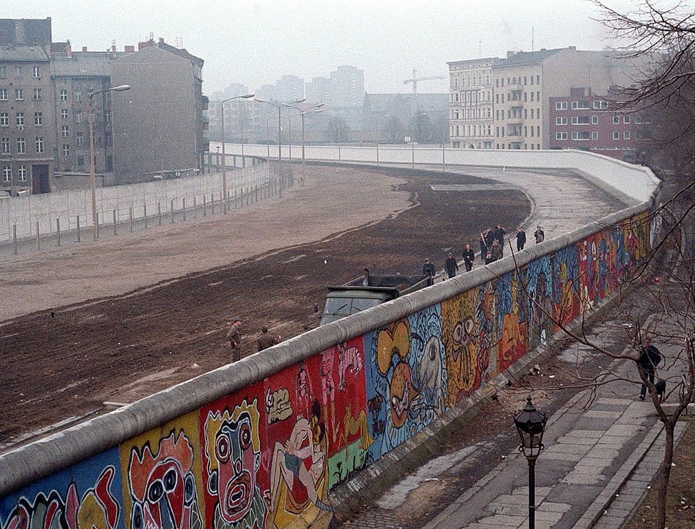 Photo of a wall dividing a city, brightly painted with graffiti on the western side, an open killing field on the eastern