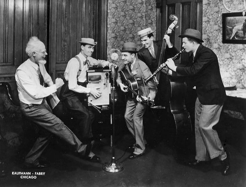 B&W photo of a quartet of musicians posing as if playing before a radio microphone, a bearded man is dancing on the left