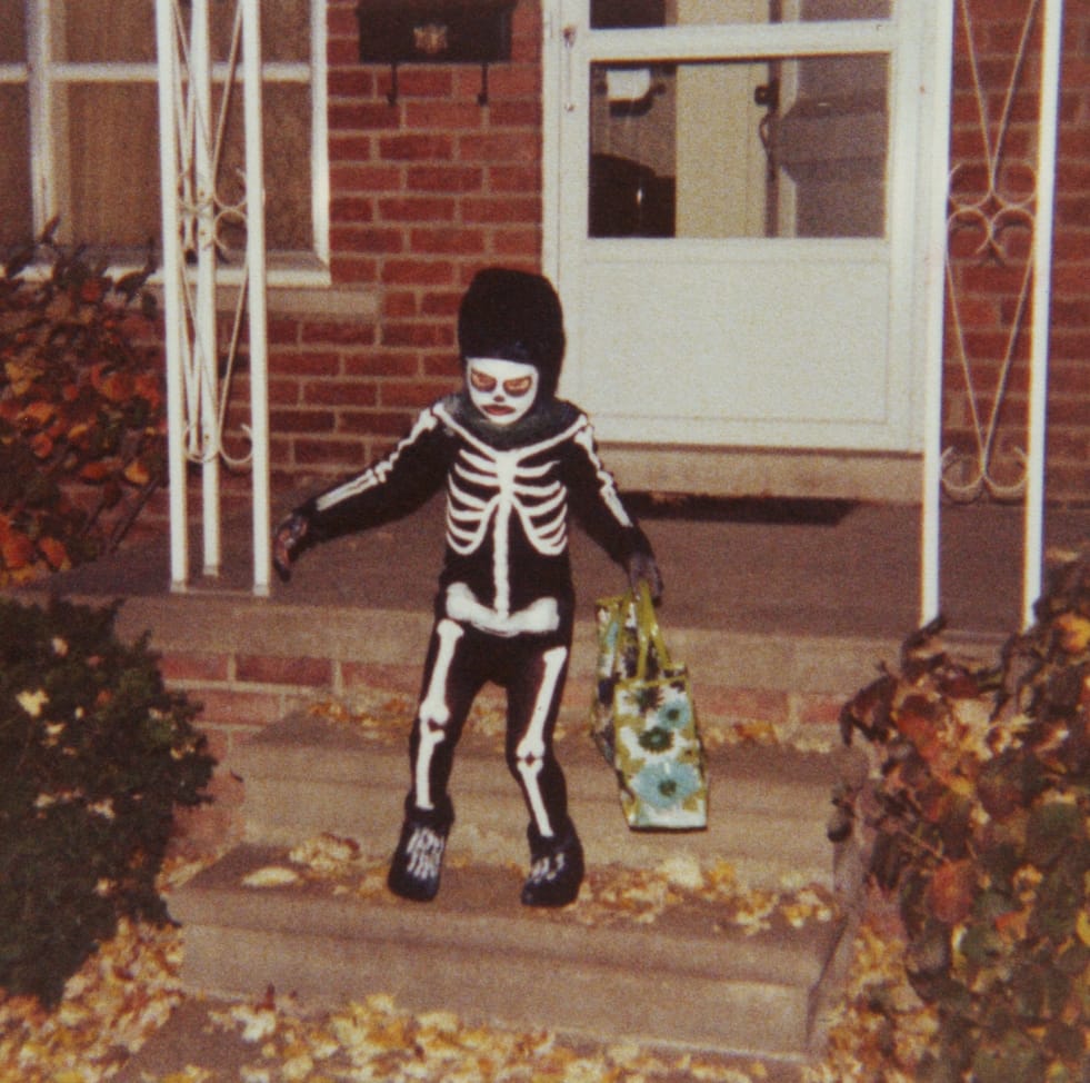 Photo of a child dressed in a skeleton costume leaving the front porch of a house carrying a shopping bag filled with candy