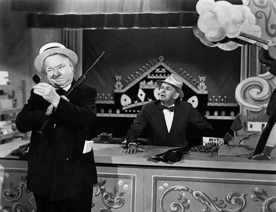 B&W photo of W. C. Fields trying to win a cigar at a shooting gallery by using a mirror to fire the rifle over his shoulder