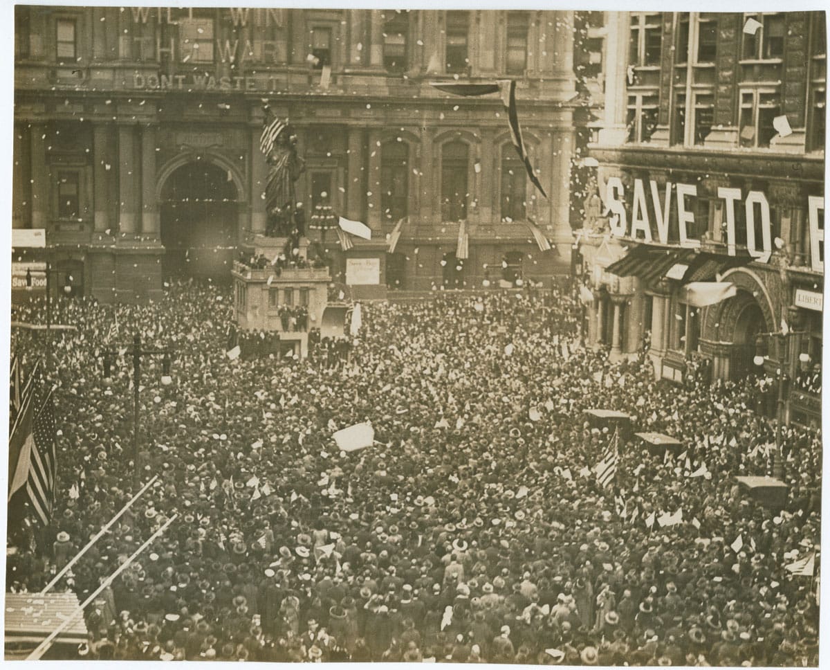 B&W photo of a crowd gathered in a city square, waving American flags and throwing confetti