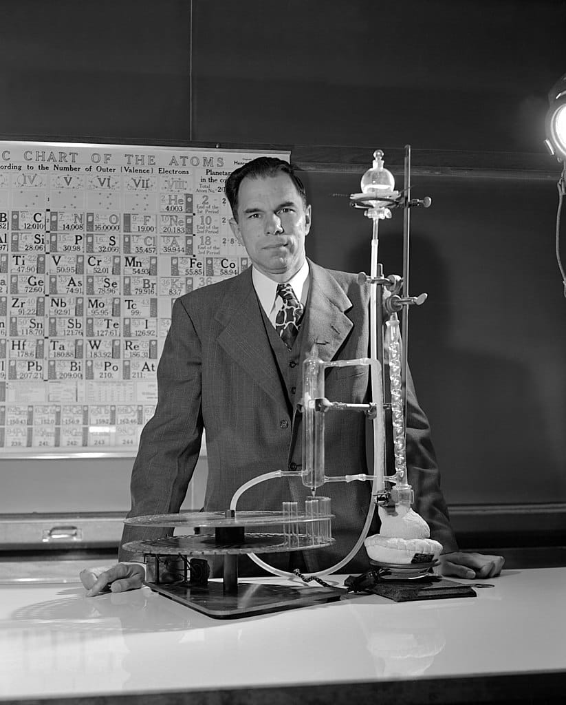 B&W photo of a man in a suit standing behind a lab bench with chemical equipment. A periodic table hangs on the wall behind.