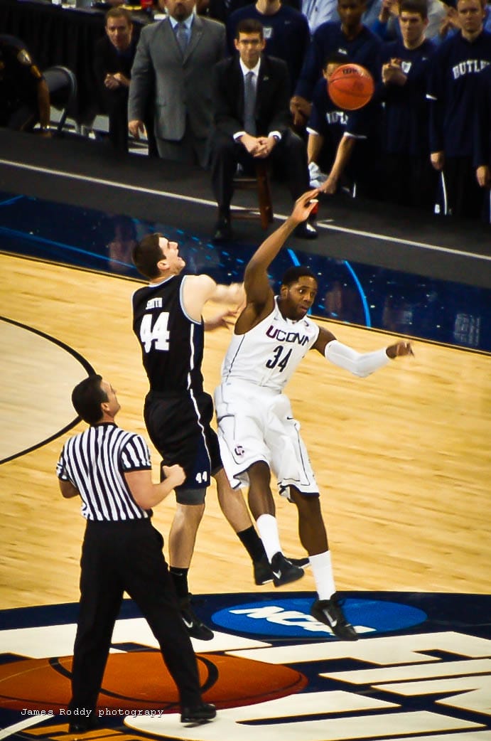 Two basketball players leap to get the opening tip of a match; a referee looks on