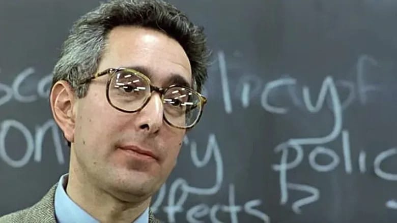 Headshot of a man (Ben Stein) standing before a blackboard with economic terms written on it