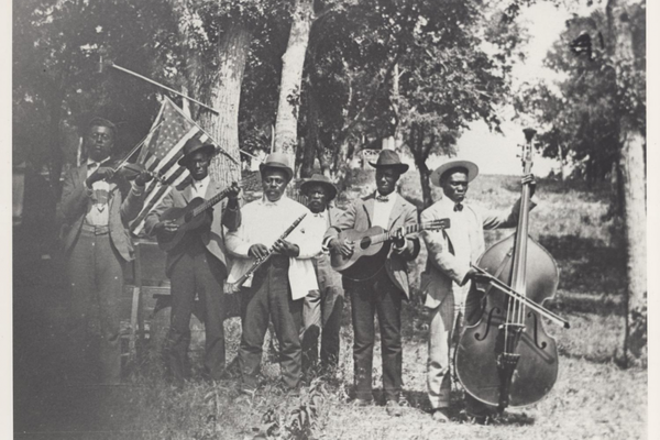 Black-and-white photo of seven Black men with musical instruments standing outdoors; an American flag is in the background
