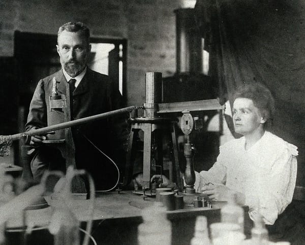 B&W photo of a man and a woman in early 20th-century dress. The woman is seated and measuring something on a scale. 