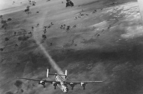 Black-and-white photo of an American B-24 bomber emerging from a flak barrage with one engine on fire