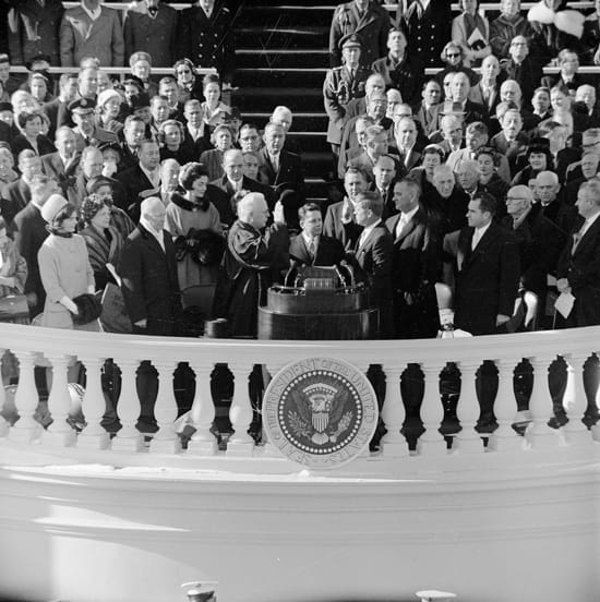 B&W photo of JFK taking the oath of office from Chief Justice Earl Warren, onlookers include Eisenhower, Nixon, and Johnson