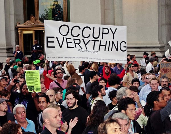 A crowd of protesters in front of a large, granite building, with two holding up a sign reading “Occupy Everything”