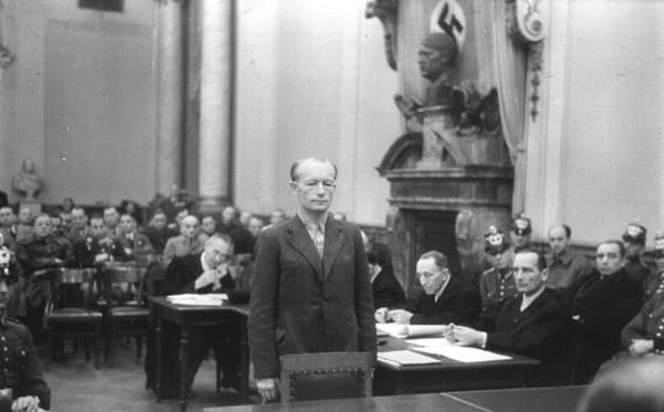 B&W photo of a man in a suit standing before a court; a Nazi flag and bust of Hitler is in the background