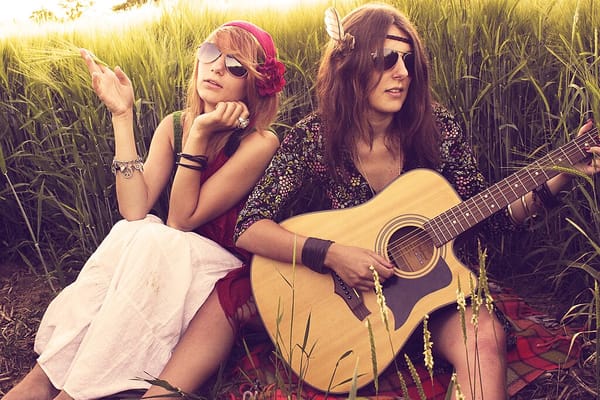 Two women in sunglasses and dressed as hippies, one holding a guitar, sitting in tall grass