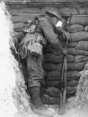 B&W photo of a WWI soldier with a rifle standing in a trench and looking through a gap in a wall of sandbags